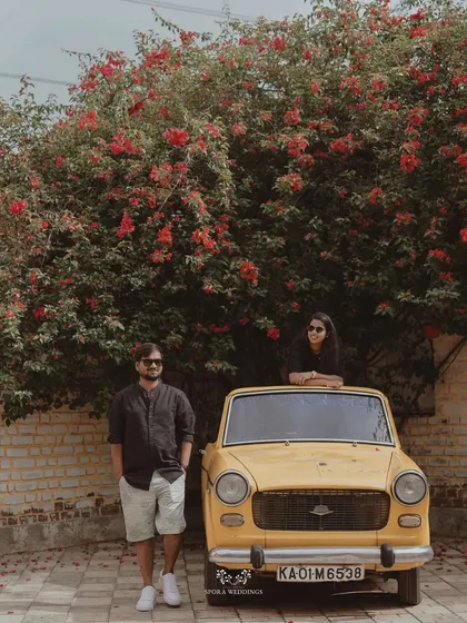The couple posing with a vintage car under a vibrant bougainvillea tree, a picture of retro romance.