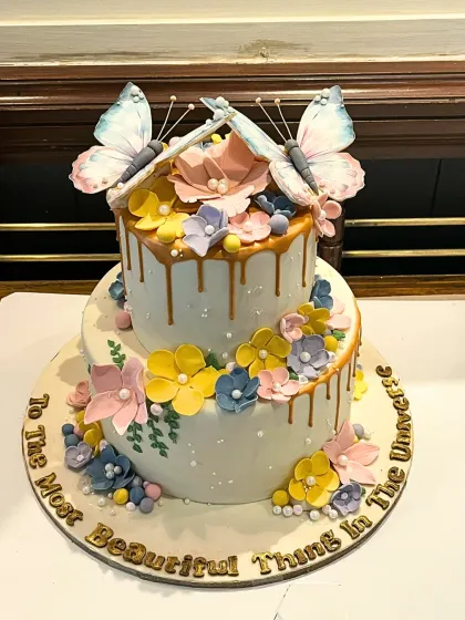A two-tier butterfly garden cake with a golden drip, adorned with colorful sugar flowers and delicate, edible butterflies.