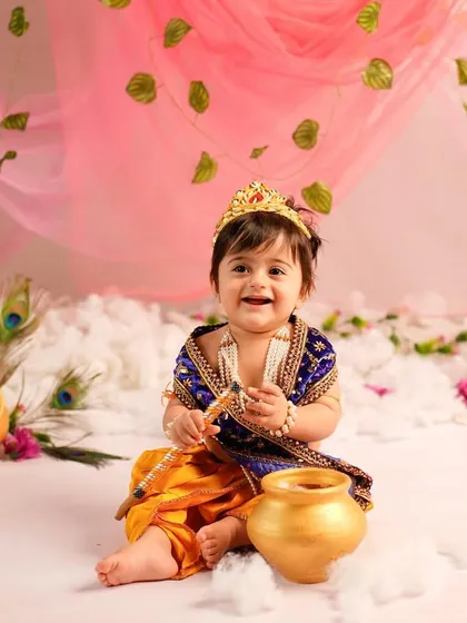 A joyful little Krishna, dressed in a beautiful blue and yellow outfit, shares a happy grin. The set is decorated with peacock feathers and golden pots for an authentic festive feel.
