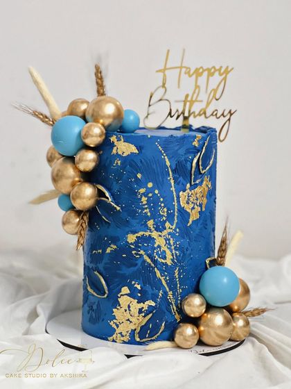 A tall, elegant chocolate ganache cake in a deep royal blue. The abstract gold leaf and texture, combined with a cluster of blue and gold spheres, create a luxurious look.