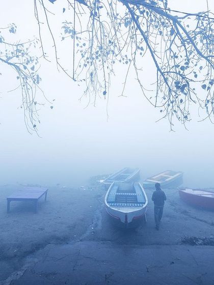 A lone figure walks by boats on the shore of the Yamuna, the bare branches of a tree framing the minimalist and serene foggy scene.