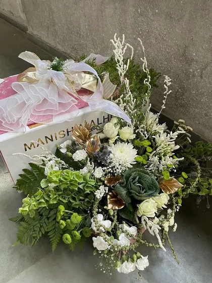 A luxurious gift arrangement featuring a designer shopping bag from Manish Malhotra, overflowing with a lavish bouquet of white and green florals. This is the epitome of high-end collaborative gifting.