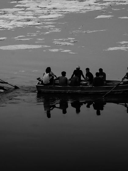 A stark black and white image showing a boat on the polluted Yamuna river, the floating foam creating a textured, abstract surface that tells a story of environmental impact.
