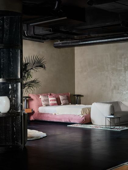 A bedroom setting featuring a low, plush pink bed. The dark, exposed ceiling and industrial pipes contrast with the soft, luxurious feel of the furniture.