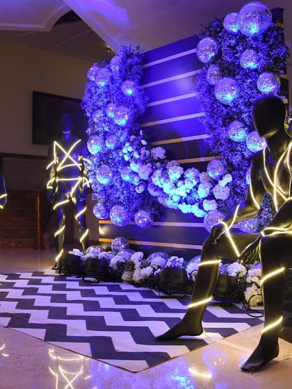 A modern and artistic installation for a Sangeet. Illuminated mannequins are placed in a lounge area with a disco ball floral wall and a chevron-patterned floor, creating a unique and edgy vibe.