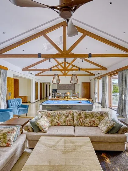 The great room of the Kabini farmhouse, designed for leisure and social gatherings. A high, vaulted ceiling with exposed wooden beams provides a sense of rustic grandeur, while large glass doors offer uninterrupted views of the pool and surrounding landscape.