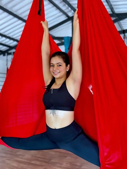 A student smiles as she opens her hips in a wide-legged split supported by the red aerial silk. Our courses are designed to be empowering and joyful.