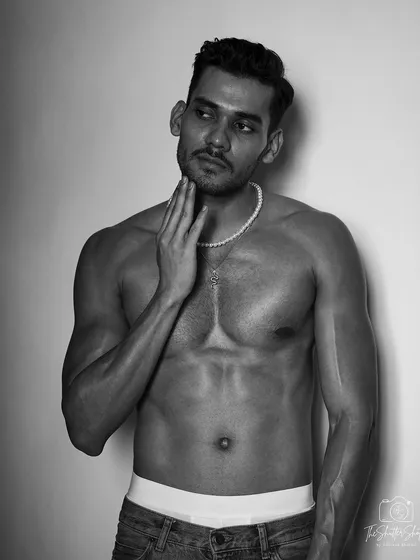 A black and white studio shot focusing on the model's upper body definition. The pose, with a hand to the neck, adds a touch of fashion to a classic physique photo.