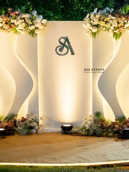A modern and minimalist Sangeet backdrop. The design features clean, curving lines with integrated backlighting and a custom couple's monogram, topped with lush floral arrangements for a touch of organic elegance.