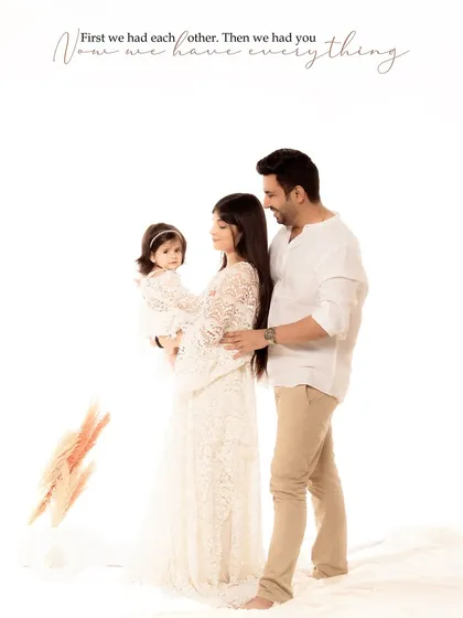 A family of three, dressed in elegant white and neutral tones, stands together in a minimalist studio setting. The text overlay, "First we had each other. Then we had you. Now we have everything," perfectly frames this love story.