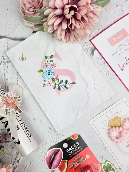 A flat lay of a bridesmaid proposal box. It includes a personalized marble-print diary, a "Bride Tribe" survival kit, lip balm, and a "Will you be my bridesmaid?" card.
