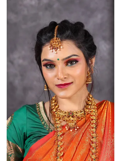 A close-up portrait of a model in a vibrant orange and green saree. The shot is framed to highlight the traditional gold jewelry and the colorful makeup.
