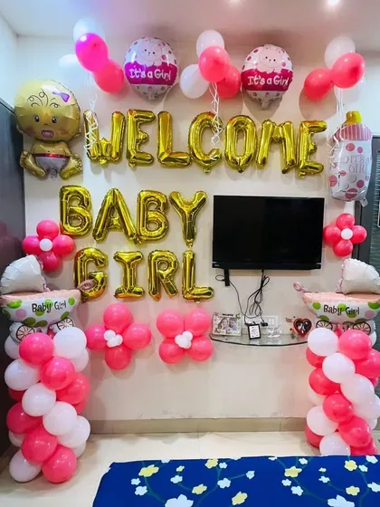 A cheerful "Welcome Baby Girl" room decoration. The wall is decorated with pink and white balloons, flower clusters, and foil balloons spelling out the welcome message.