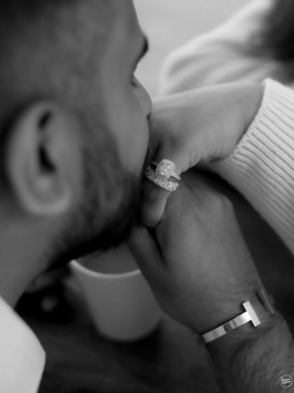 A tender, intimate black and white portrait focusing on a gentle kiss on the hand. This close-up shot highlights the beautiful engagement ring and the quiet affection between the couple.