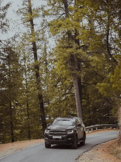 A black Ford Endeavour emerges from a curve on a road flanked by dense green forests. This image captures the feeling of exploring hidden paths and the vehicle's commanding road presence.