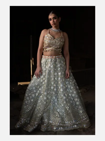 A model stands in a softly lit, dark environment, wearing a stunning silver and gold embellished lehenga. The dramatic lighting makes the outfit sparkle, emphasizing its luxurious quality for evening or bridal wear.