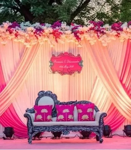 A beautiful reception stage with a backdrop of pink and cream drapes. The vintage-style sofa with bright pink cushions adds a pop of color and personality.