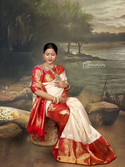 A serene studio portrait of a mother-to-be in a traditional red and white saree, seated against a painted backdrop.