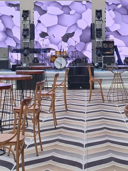 A modern stage setup for a corporate event. The backdrop features a geometric, backlit LED screen, with rustic high-top tables and chairs in the foreground for a relaxed, contemporary vibe.