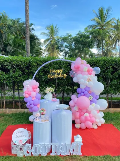 Our decorations are perfect for outdoor celebrations too. Here is a vibrant birthday setup with a circular arch, a colorful balloon arrangement in pink and purple, and personalized name blocks, all arranged for a garden party.