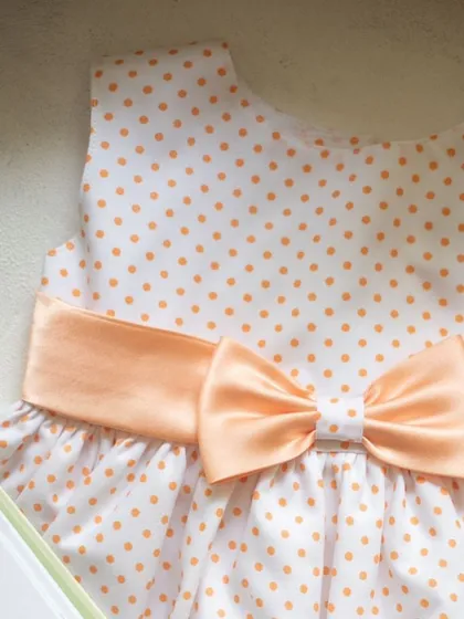 A classic combination of peach polka dots on a white background. The wide satin bow at the waist adds a touch of elegance and defines the silhouette beautifully.