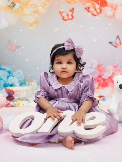 A sweet portrait of a one-year-old girl in a purple dress, holding a "one" sign in a whimsical, colorful setup.