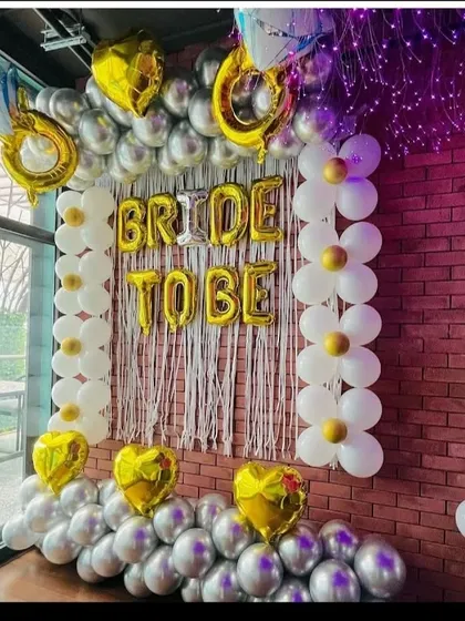 A fun 'Bride to Be' decoration against a brick wall. This setup uses gold foil letters, a white fringe curtain, and columns of daisy-shaped balloons made from white and gold balloons.