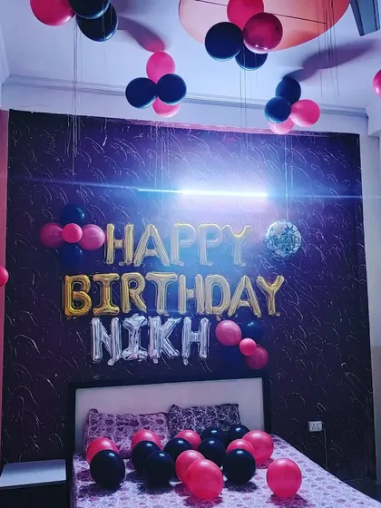 A birthday room decoration with a red and black color scheme. The setup includes 'Happy Birthday' foil balloons with a custom name, and balloon bunches hanging from the ceiling.