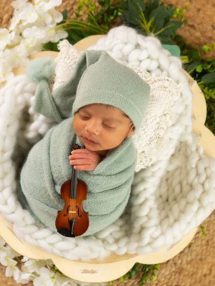 A newborn in a sage green wrap and hat holds a miniature violin, nestled in a soft white basket surrounded by greenery.
