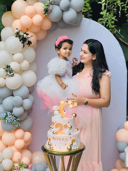 Diyara celebrating her 2nd birthday with her mom. The backdrop features a gentle arch with a beautiful arrangement of pastel peach and grey balloons, creating a soft and elegant look.