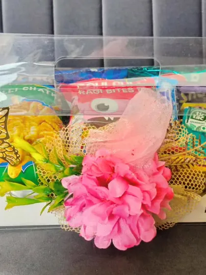 For a budget-friendly option, I offer these lovely welcome hampers in a clear box. They are filled with a variety of snacks and decorated with a bright pink flower and gold butterflies for a cheerful touch.