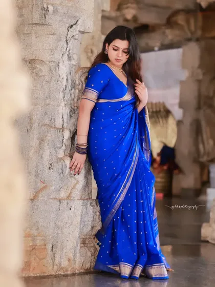 A full-length portrait showing the beautiful drape of the blue saree against a stone wall.