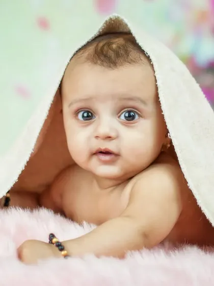 A stunning close-up of a baby with beautiful, wide eyes, peeking out from under a towel. The soft, colorful background adds a dreamy quality to this captivating portrait.