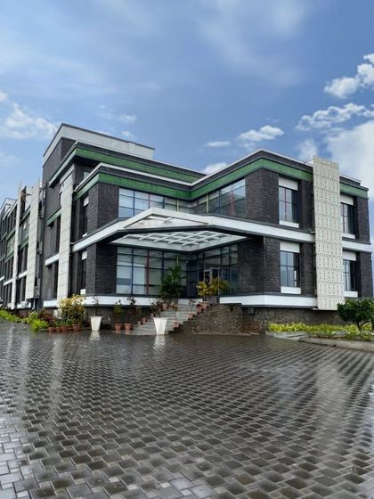 The entrance of Venkateshwar World School in Pune after a rain shower. The wet cobblestones reflect the building's facade, highlighting the material palette of grey stone, white GRC jalis, and green accents.