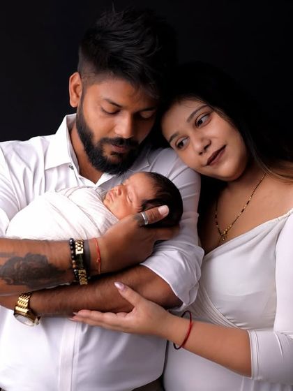A complete family portrait with their newborn. The parents gaze down at their baby, wrapped snugly in white, creating a timeless image of love and unity against a classic black studio backdrop.