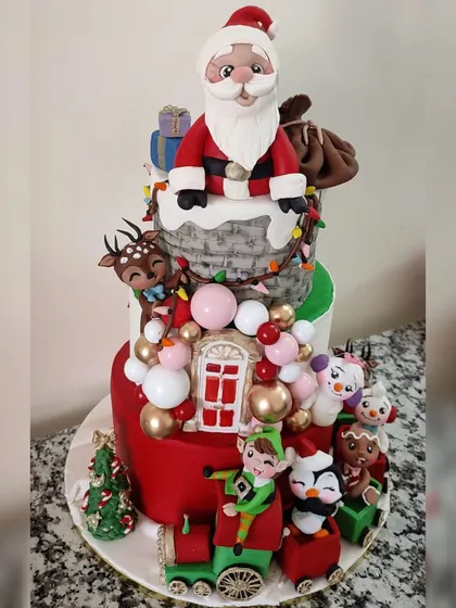 A full view of the incredible three-tiered Christmas cake, packed with numerous handcrafted figurines. It features Santa, an elf, a reindeer, snowmen, a gingerbread man, and a penguin, all riding a festive train.
