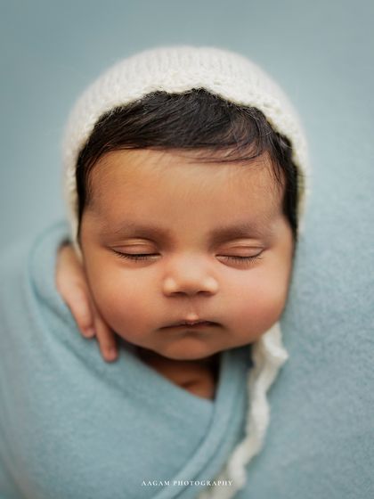Those squishy cheeks are just irresistible. A close-up portrait of a newborn in a simple white bonnet against a soft blue background.