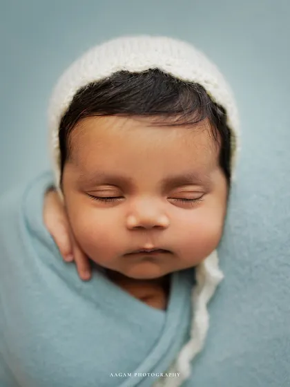 Those squishy cheeks are just irresistible. A close-up portrait of a newborn in a simple white bonnet against a soft blue background.