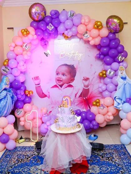A princess-themed first birthday featuring a beautiful photo of the birthday girl. The decor is set with a purple and pink balloon circle, character cutouts, and a neon number '1'.