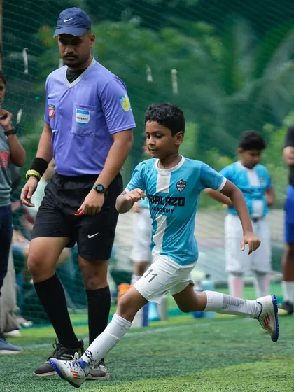 A young player on the run, focused on the ball with a referee close by, during an intense moment in a grassroots match.