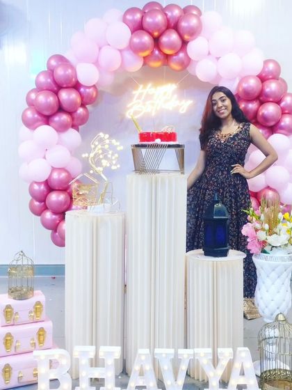 The birthday girl posing with her beautiful pink-themed balloon arch decoration. The setup includes multiple plinths for the cake and gifts.