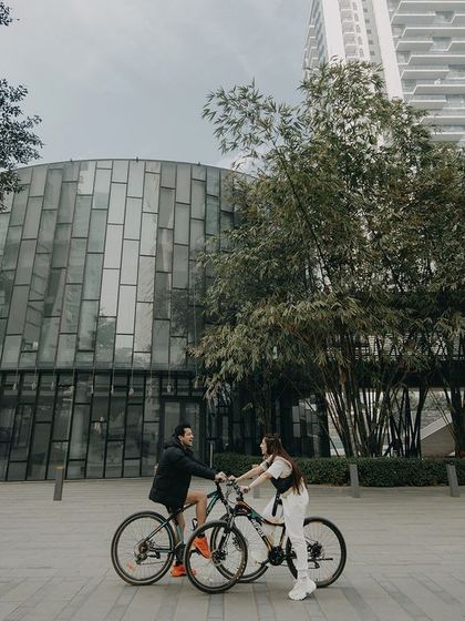 A wide shot capturing a couple's cycling adventure against modern architecture. This pre-wedding photo tells a story of shared activities and urban exploration.