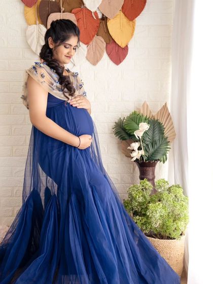 A serene solo portrait of a mom-to-be in a blue gown, standing in a naturally lit studio with bohemian decor.