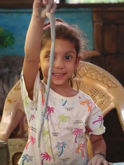 A young student proudly holds up a long coil of clay, ready to start building her pot. We teach children fun and accessible techniques to help them create with confidence.