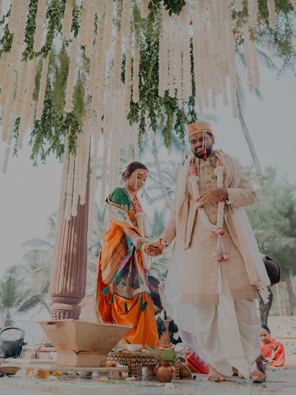 A beautiful, full-length shot of the couple taking their pheras around the sacred fire in a traditional Maharashtrian wedding, framed by hanging floral decor.