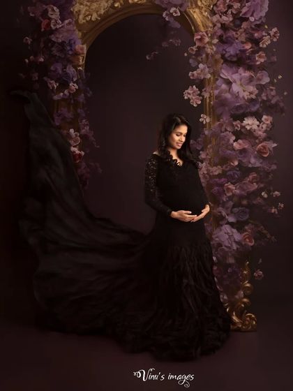 A portrait of dark elegance. The mother-to-be, in a stunning black lace and tulle gown, stands within a frame of purple flowers, creating a moody and romantic atmosphere.