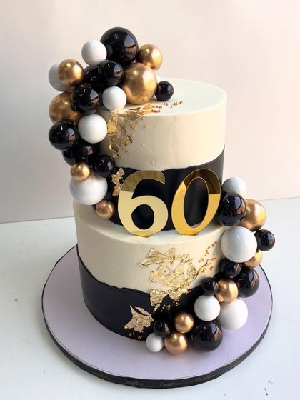 Black, white, and gold perfection. This classy two-tiered cake for a 60th celebration is finished with luxe metallic spheres, delicate gold leaf details, and a bold topper, designed to steal the spotlight.