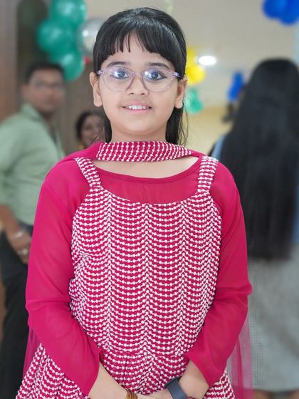 A portrait of a smiling young girl with glasses, dressed in a pink and white outfit. We capture the unique charm of every child we photograph.