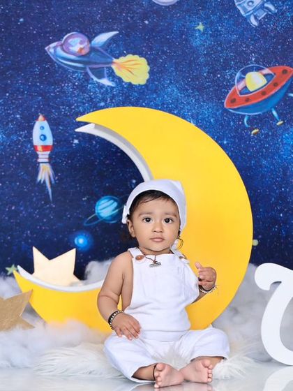 A baby boy in a white astronaut outfit sits among the stars and clouds on a crescent moon. The "ONE" sign makes this a perfect theme for a first birthday adventure.
