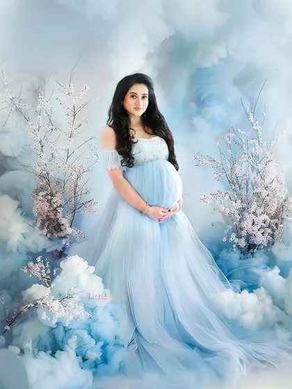 A dreamy and ethereal solo maternity portrait. The expecting mother is dressed in a flowing light blue gown, surrounded by a magical set of soft clouds and delicate white blossoms, creating a fantasy-like atmosphere.